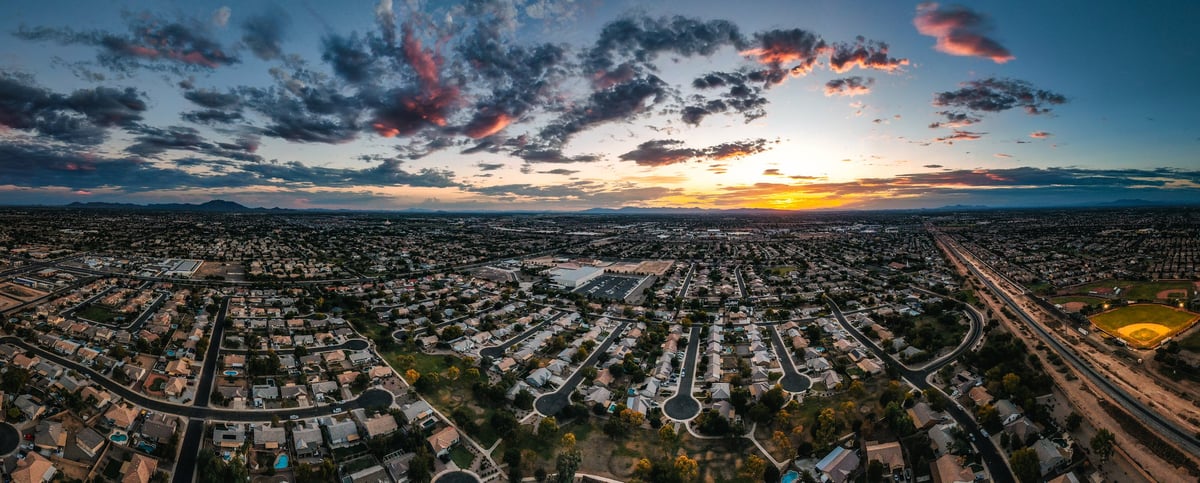 Aerial panorama over a Phoenix suburban neighborhood at dusk — tile rooftops stretch across the valley with desert mountains silhouetted on the horizon