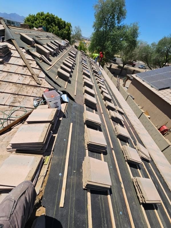 Roofing installation in progress with workers on a steep residential roof surrounded by trees and materials